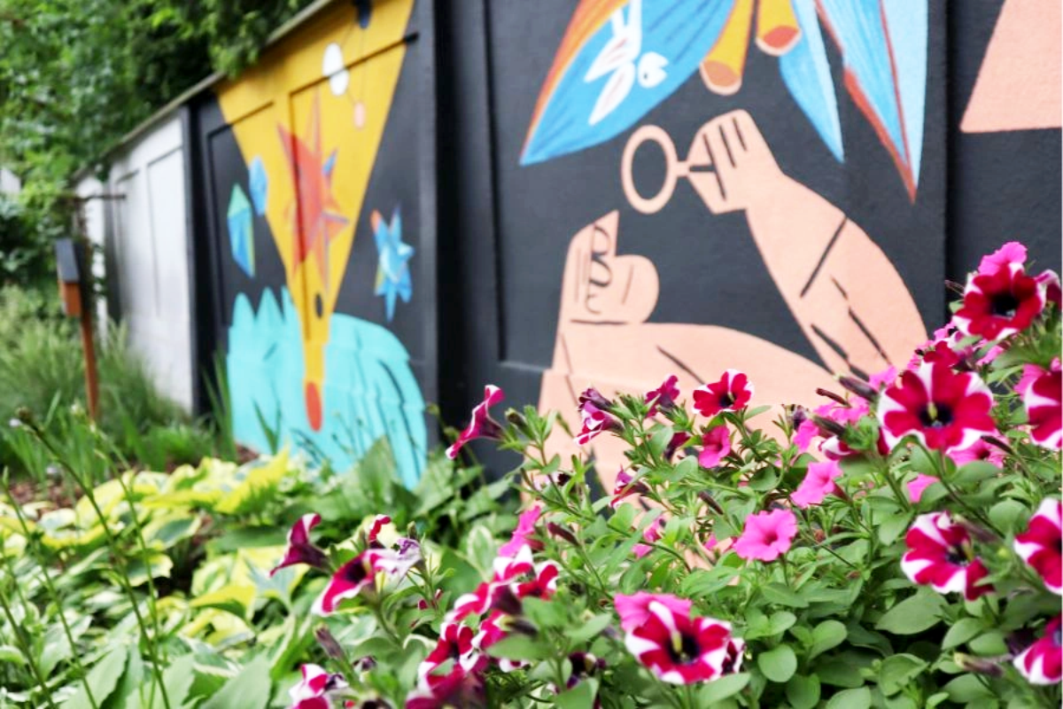 Kolorowy mural na czarnej ścianie z abstrakcyjnymi wzorami, takimi jak tęcza, dłoń trzymająca lupę i geometryczne kształty w żywych barwach, w pierwszym planie kwitnące różowo-fioletowe petunie i zielone liście roślin.